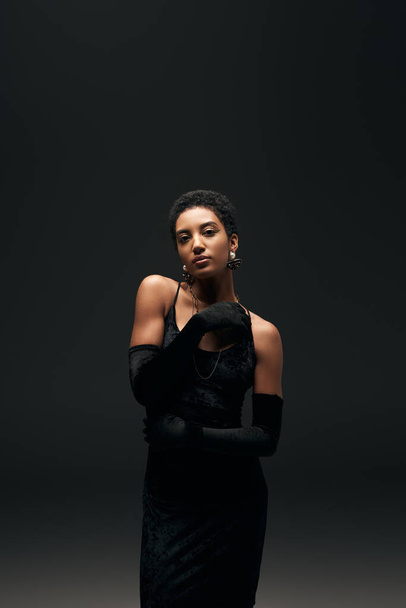 Elegant african american woman in gloves, golden necklaces and little dress looking at camera while standing isolated on black, high fashion and evening look - Fotoğraf, Görsel