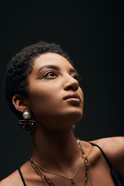 Portrait of elegant and short haired african american woman with evening makeup and golden accessories looking away and standing isolated on black, high fashion and evening look - Fotografie, Obrázek