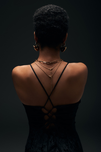 Vista posterior de la mujer afroamericana de moda con pelo corto y accesorios de oro de pie aislado en negro con iluminación, alta moda y aspecto de noche, joyas, femenino, sensualidad  - Foto, Imagen