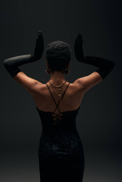 Back view of elegant african american woman in evening dress, gloves and golden accessories posing isolated on black with lighting, high fashion and evening look, feminine sensuality  - Foto, Imagem