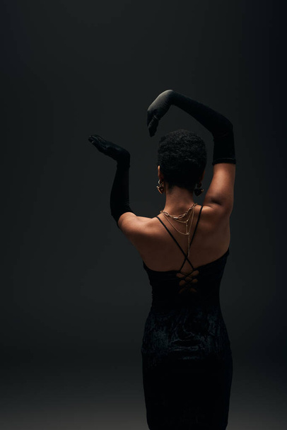 Vista posterior de la mujer afroamericana elegante y de pelo corto en guantes, vestido de noche y accesorios dorados posando aislados en negro, alta moda y aspecto de noche - Foto, Imagen