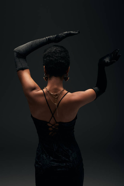 Back view of fashionable african american woman in gloves, golden accessories and evening dress posing while standing isolated on black, high fashion and evening look - Φωτογραφία, εικόνα