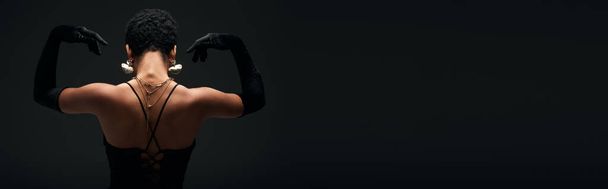 Vista posteriore della giovane donna afroamericana alla moda in guanti e accessori dorati in posa in illuminazione mentre in piedi isolato su nero, alta moda e look da sera, banner  - Foto, immagini