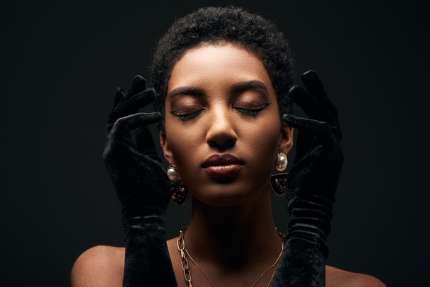 Portrait of elegant and short haired african american woman with evening makeup, golden chains, earrings and gloves posing with closed eyes isolated on black, high fashion and evening look - Foto, afbeelding
