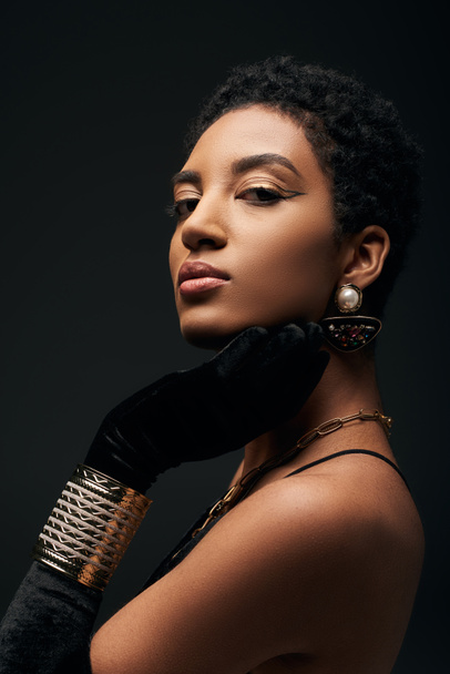 Portrait of confident and elegant young african american woman with evening makeup, glove and golden accessories looking at camera isolated on black, high fashion and evening look - Fotó, kép