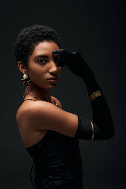 Fashionable and short haired african american model in dress, accessories and glove covering eye while posing isolated on black, high fashion and evening look - Foto, Imagem