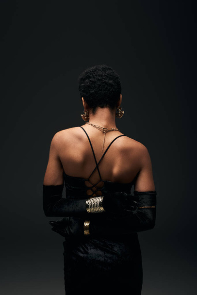 Widok z tyłu eleganckie i krótkie włosy Afroamerykanka w rękawiczkach, sukienka i nowoczesne akcesoria stojące odizolowane na czarny, wysoka moda i wieczorem wygląd, kobiecy - Zdjęcie, obraz