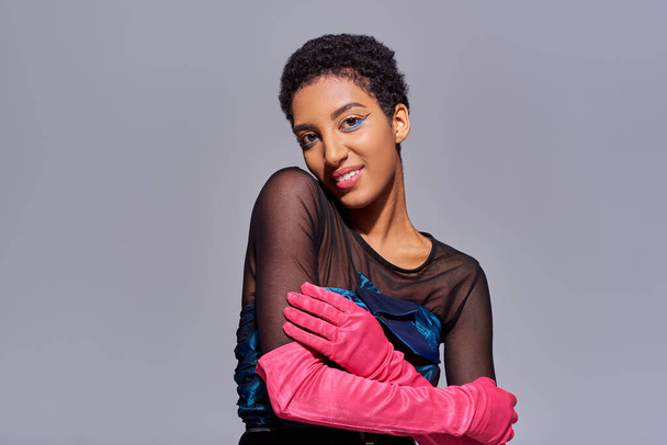 Uśmiechnięta i modna afrykańska modelka w sukience koktajlowej i różowych rękawiczkach dotykająca ramienia i patrząca w kamerę podczas pozowania na szarej, nowoczesnej generacji koncepcji mody z - Zdjęcie, obraz