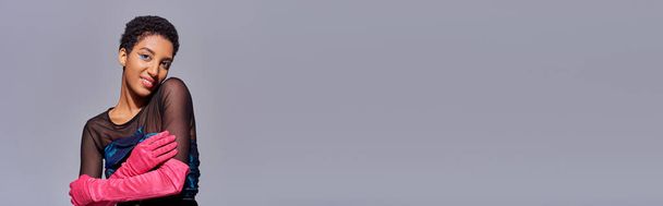 Modelo americano africano de cabelos curtos e positivos em vestido de cocktail e luvas rosa tocando braço e de pé isolado no conceito de moda z de geração moderna, cinza, banner com espaço de cópia - Foto, Imagem