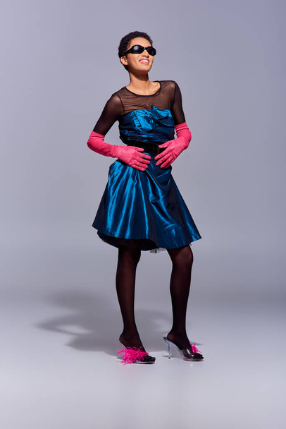 Full length of cheerful and stylish african american woman in sunglasses, cocktail dress and heels with feathers posing on grey background, modern generation z fashion concept - Zdjęcie, obraz