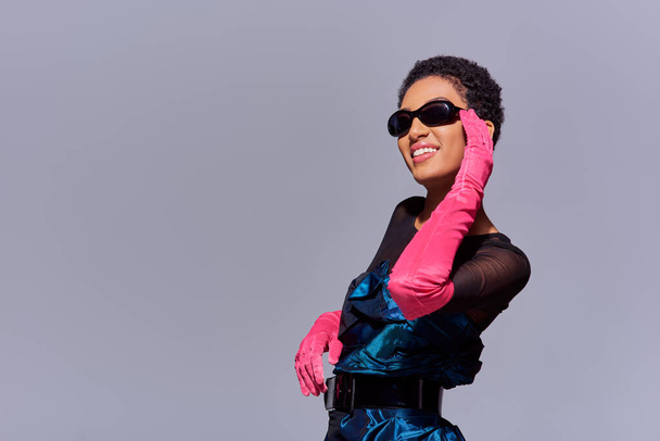 Fröhliche afrikanisch-amerikanische Frau in Cocktailkleid und rosa Handschuhen, die Sonnenbrille berührt und isoliert auf grauem, modernem Modekonzept der Generation Z steht - Foto, Bild