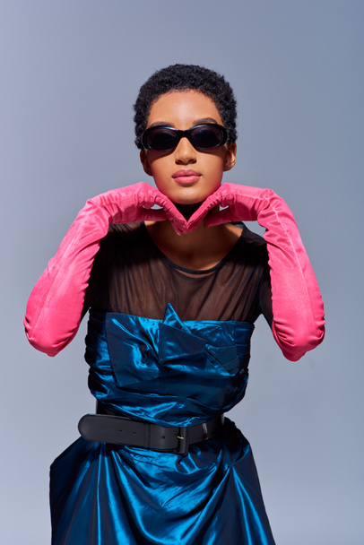 Αυτοπεποίθηση νεαρή Αφροαμερικανή γυναίκα σε γυαλιά ηλίου, ροζ γάντια και κοκτέιλ φόρεμα αγγίζοντας το πηγούνι, ενώ στέκεται απομονωμένη σε γκρι, μοντέρνα γενιά z έννοια της μόδας - Φωτογραφία, εικόνα