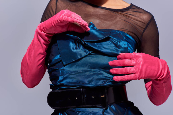 Cropped view of young model in stylish cocktail dress and pink gloves touching bow while standing isolated on grey, modern generation z fashion concept, details, belt, feminine - Photo, Image