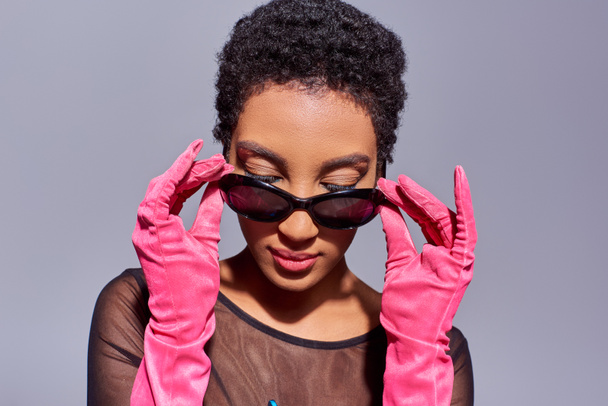 Молода і впевнена афро-американська жінка торкається сонцезахисних окулярів і одягає рожеві рукавички, виставляючи і стоячи ізольовано на сірому фоні сучасного покоління z-концепції - Фото, зображення