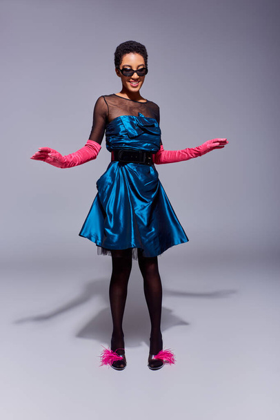 Full length of cheerful african american woman in sunglasses, cocktail dress and pink gloves posing in feathered shoes while standing on grey background, modern generation z fashion concept - Foto, Bild