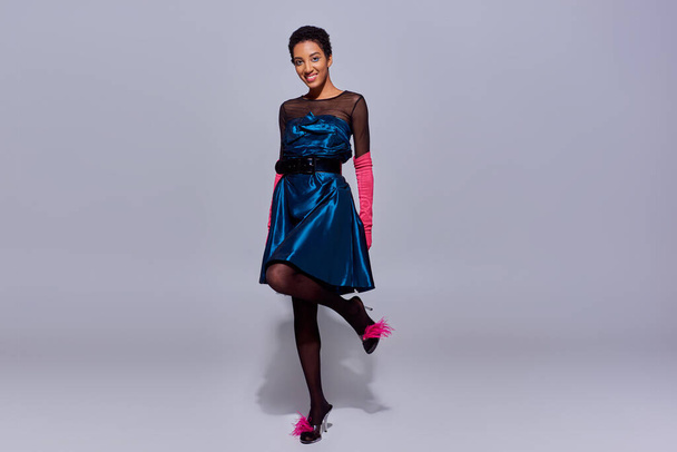 Longitud completa de la mujer afroamericana positiva en guantes de color rosa, vestido de cóctel y zapatos emplumados posando y de pie sobre fondo gris, concepto de moda z generación moderna - Foto, Imagen