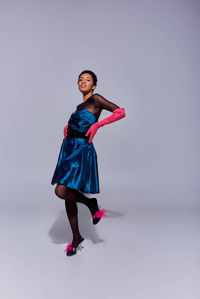 Trendy african american woman in cocktail dress, pink gloves and heels with feathers touching hips and smiling while standing on grey background, modern generation z fashion concept - Photo, Image