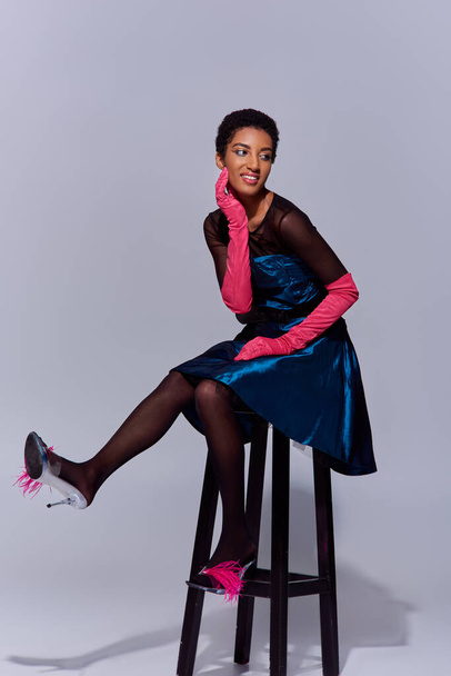 Vreugdevol en stijlvol Afrikaans Amerikaans model in roze handschoenen, cocktailjurk en hakken met veren poserend op stoel op grijze achtergrond, modern generatie z modeconcept - Foto, afbeelding
