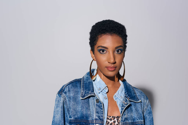 Glamorous african american woman with bold makeup and golden earrings wearing denim jacket and looking at camera while standing on grey background, denim fashion concept - Photo, Image