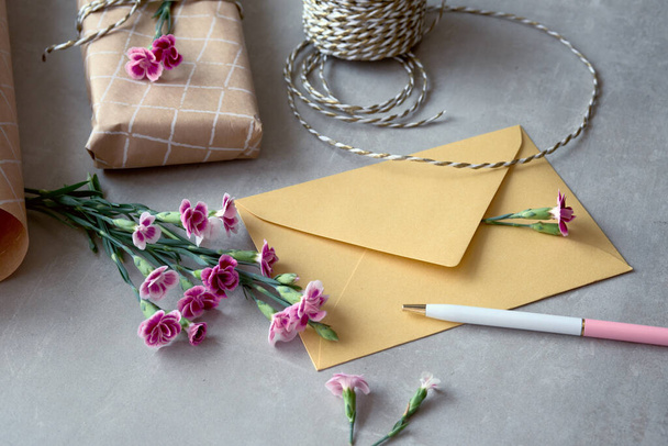 Envelope and self made greeting card concept with fresh carnation flowers. Wrapped gift, paper postcard, paper envelope, cord, scissors and purple carnations. - Foto, immagini