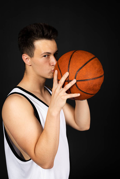 Handsome young basketball player in white sportswear kisses a ball. Isolated on black background. view from behind - Fotografie, Obrázek