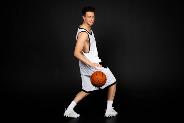 Όμορφος νεαρός μπασκετμπολίστας με άσπρα αθλητικά κρατά μια μπάλα. Απομονωμένο σε μαύρο φόντο. θέα από πίσω - Φωτογραφία, εικόνα