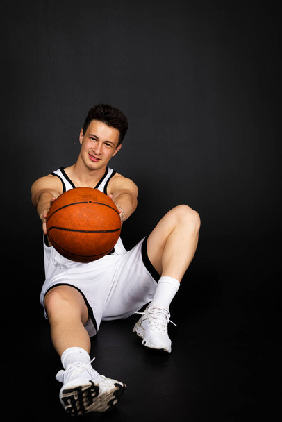 白いスポーツウェアを着たハンサムな若いバスケットボール選手がボールでポーズをとって床に座っています。黒の背景に隔離された. - 写真・画像