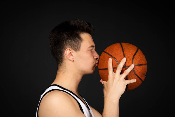 Handsome young basketball player in white sportswear kisses a ball. Isolated on black background. view from behind - Foto, afbeelding