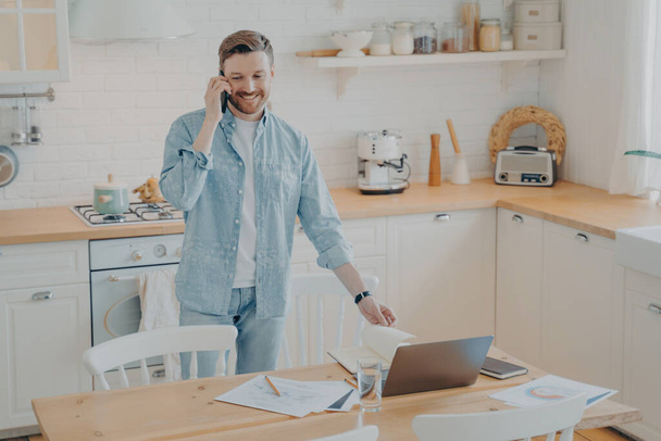 Casual geklede man multitasking aan keukentafel. Werken aan een laptop, telefoneren met een vriend. Vrijwilligerswerk en telewerk omarmen. - Foto, afbeelding