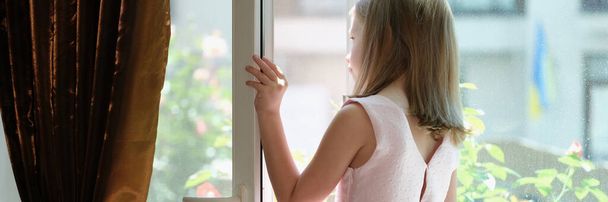 Close-up of little girl in pink dress standing on windowsill at open window. Danger of falling from height concept - Fotoğraf, Görsel