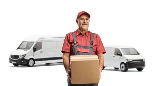 Cheerful mature delivery man in front of transport vans carrying a cardboard box isolated on white background - Foto, immagini