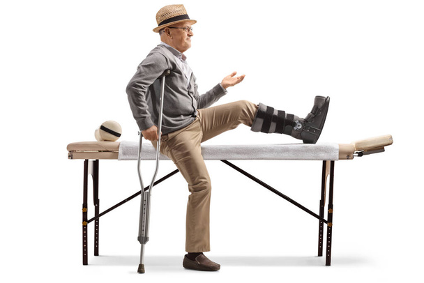 Senior man with a leg brace sitting on a bed for physical therapy isolated on white background - Photo, image