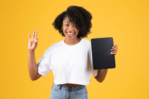 Θετική χιλιετή αφροαμερικάνικη σγουρή γυναίκα δείχνουν tablet με λευκή οθόνη, κάνουν ok χειρονομία, απομονώνονται σε κίτρινο φόντο. Σύσταση και έγκριση, διαφήμιση και προσφορά, εφαρμογή κοινωνικής δικτύωσης και blog - Φωτογραφία, εικόνα