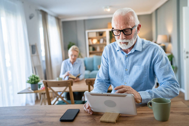 Homme âgé caucasien vieux grand-père mâle assis à la maison utiliser une tablette numérique pour l'application d'appel vidéo en ligne parler tandis que la femme s'asseoir derrière des personnes réelles hors foyer espace de copie concept de loisirs famille - Photo, image