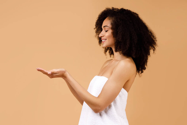 Mujer negra joven envuelta en toalla de baño demostrando algo en las palmas vacías, mostrando objetos invisibles y sonriendo, posando sobre fondo beige del estudio, vista lateral, espacio para copiar - Foto, Imagen
