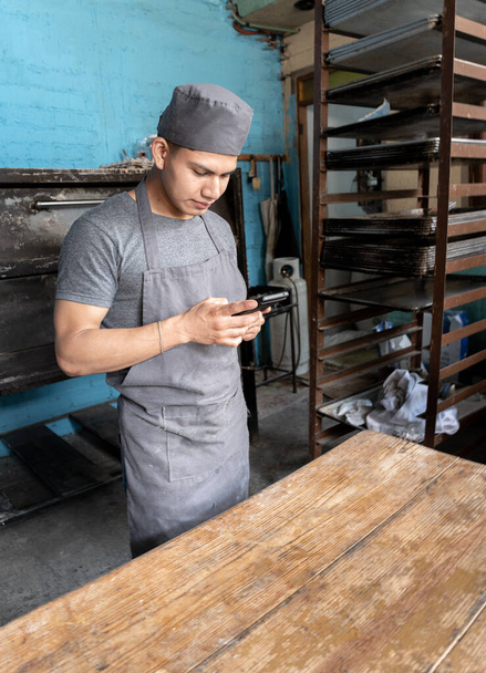 Um jovem padeiro hispânico está a fazer uma pausa para usar o telefone na padaria. Ele está usando um avental cinza e um uniforme tradicional de padeiro. Conceito de pão doce mexicano tradicional - Foto, Imagem
