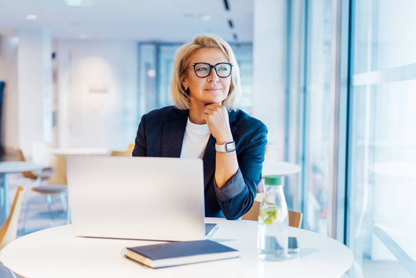 50s confident mature businesswoman dreamy looking at window, middle-aged experienced senior female professional working on laptop in open space office. Female entrepreneur working remotely - Zdjęcie, obraz