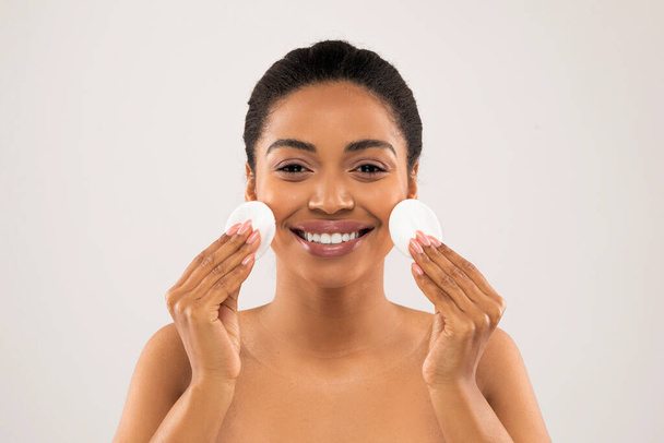 Headshot of happy smiling half-naked beautiful young african american woman holding cotton pads next to her cheeks, cleansing skin, using micellar water or toner, gray studio background - Foto, Bild