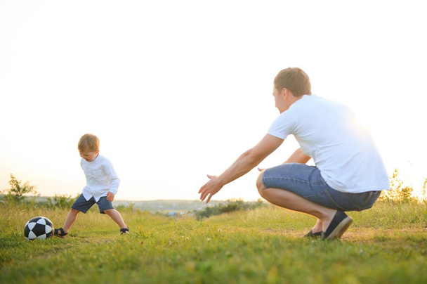 father with a little son plays football on the green grass in the park. Happy family having fun and playing football on a green grassy lawn on a sunny day. Family concept, Father's Day. - Photo, Image