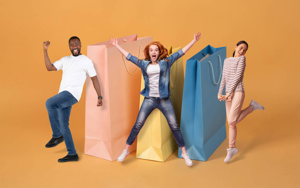 Multiracial friends emotional millennial two ladies and one guy gesturing next to huge shopping bags over colorful studio background, celebrating season sale, mockup, collage, full length - Photo, Image