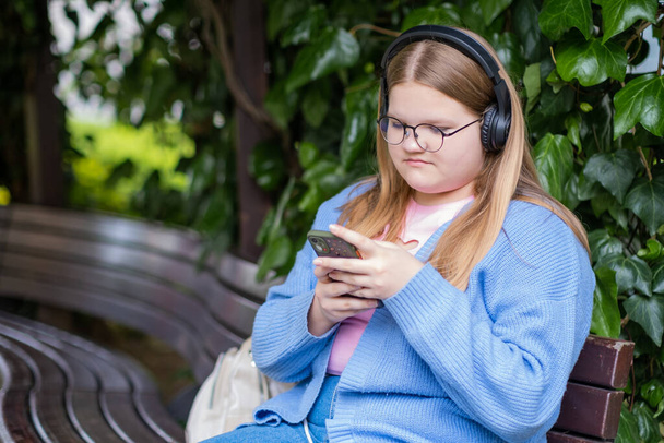 Koncentrált tinédzser vezeték nélküli fejhallgató ül a padon a Spring Park mobiltelefon szőke lány szemüvegben ellenőrzi a közösségi média keresztül modern eszköz - Fotó, kép