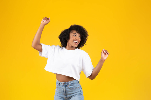 Happy millennial african american curly woman in white t-shirt has fun, dancing, looks at empty space, isolated on yellow studio background. Ad and offer, sale, lady enjoy free time, music - Fotó, kép