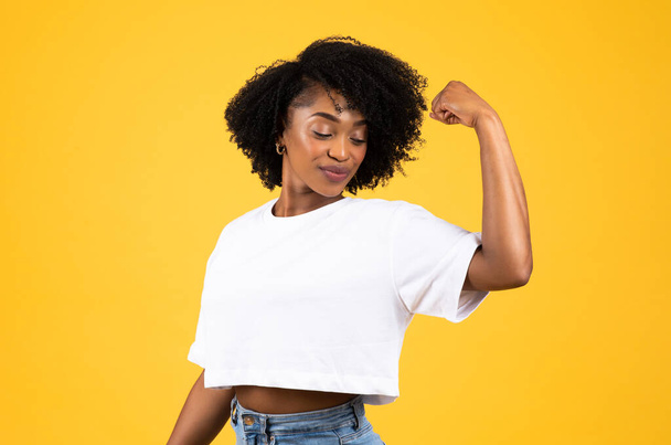 Felice forte fiducioso millenaria donna riccia africana americana in t-shirt bianca mostrano muscoli bicipiti, isolati su sfondo giallo studio. Stile di vita, forza, potenza, annuncio e offerta - Foto, immagini