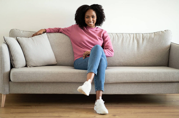 Sorrindo confiante millennial afro-americano encaracolado senhora em casual sentado no sofá, desfrutar de conforto e tempo livre na sala de estar interior. Descanso, relaxamento, pausa e estilo de vida em casa - Foto, Imagem