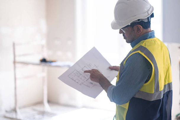 Specialist examining interior project drawing standing against construction safety equipment man in professional uniform stands in room controlling renovation process in apartment - Photo, Image