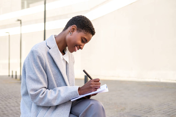 retrato horizontal de uma jovem mulher africana atraente. Ela está sentada perto do prédio de escritórios e escrevendo em um caderno de papel - Foto, Imagem