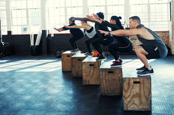 Estirarse lo más lejos posible. un grupo enfocado de jóvenes que se abalanzan sobre cajas como ejercicio dentro de un gimnasio - Foto, imagen