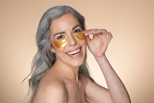 Ritratto di felice bella donna anziana dai capelli grigi che indossa macchie d'oro sotto gli occhi, si prende cura della sua pelle, sorridendo alla macchina fotografica su sfondo beige studio, spazio libero - Foto, immagini