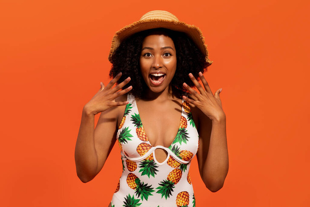 Amazed emotional thrilled beautiful curly young pretty black lady wearing wicker hat and colorful swimsuit gesturing and exclaiming over orange stuio background. Best travel offer - Photo, Image