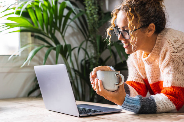 Современная женщина улыбается дисплею ноутбука и наслаждается временем в Интернете в одиночестве. Концепция коммуникации видеозвонков. Счастливые женщины используют ноутбук дома на столе и держа чашку чая - Фото, изображение